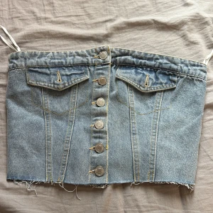Jeans tubtopp med knappar, strl S - Säljer en snygg tubtopp i ljusblå denim med knappar framtill och två fejkfickor. Toppen har rå kant nertill och smockad rygg för skön passform. Perfekt för dig som gillar en edgy och trendig stil.