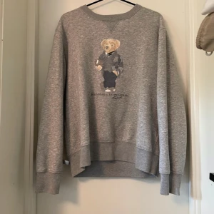 Grå sweatshirt Polo Bear Ralph Lauren - Grå sweatshirt från Polo Ralph Lauren med ikoniska Polo Bear-trycket framtill. Klassisk rund halsringning, ribbade muddar och avslappnad passform. Perfekt för dig som gillar streetstyle och vill ha en chill vibe. Materialet är mjuk bomullsblandning. Jättebra skick förutom att björnen har börjat tappa lite färg från tvättning.