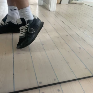 Svarta Philippe Model sneakers - Snygga svarta sneakers från Philippe Model med vita detaljer och snörning. Skorna har en klassisk låg siluett, rund tå och är tillverkade i en mix av skinn och textil. Grå sula med tydlig logga undertill och vit hälkappa för extra kontrast.