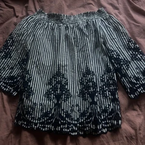 Randig offshoulder blus med brodyr - Säljer en snygg offshoulder blus med svarta och vita ränder och detaljerad svart brodyr längs ärmar och nederkant. Blusen har resår upptill och vida trekvartsärmar. Perfekt för dig som gillar unika detaljer och en luftig stil.