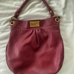 Vinröd handväska Marc by Marc Jacobs - Säljer en vinröd handväska från Marc by Marc Jacobs i mjukt skinn med guldfärgade metalldetaljer och ett stort logoplate framtill. Väskan är äkta. 