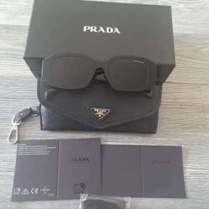Svarta solglasögon från Prada - Snygga svarta solglasögon från Prada med rektangulära bågar och breda skalmar där loggan syns tydligt. Glasögonen har en modern och edgy vibe, levereras med originalfodral och box. Perfekta för dig som vill sticka ut med en lyxig accessoar.