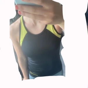 Svart och neon gul träningslinne - Sportig svart och gul träningslinne från Gymology Athletica London i storlek XS. Snygg racerback-modell med meshdetaljer i gult och tight passform. Perfekt för gymmet eller löprundan, tillverkad i stretchigt syntetmaterial som andas bra. Har S i storlek så linnet passar mycket väl storlek S.