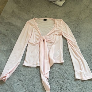 Ljusrosa knytblus från Gina Tricot - Supersöt ljusrosa blus från Gina Tricot med långa ärmar och snygg knytning framtill. Toppens material är mjukt och lätt, perfekt för en trendig look. Den har en croppad passform och v-ringad halsringning som ger en feminin vibe.