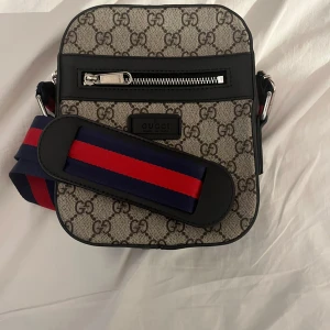 Gucci axelväska med GG-mönster - Snygg axelväska från Gucci med klassiskt GG-mönster i beige och brunt. Väskan har en bred axelrem i blått och rött, silverfärgad dragkedja framtill och svarta skinn-detaljer. Perfekt för dig som vill ha en ikonisk och trendig accessoar.