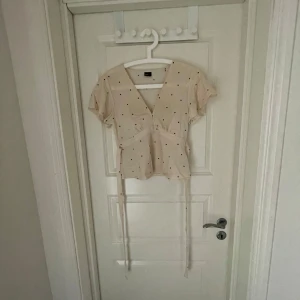 Prickig beige blus från Gina Tricot - Supersöt beige blus från Gina Tricot med svarta prickar, v-ringning och knytband i midjan. Kortärmad och lätt figursydd modell som ger en feminin siluett. Perfekt för dig som gillar en söt och trendig look.