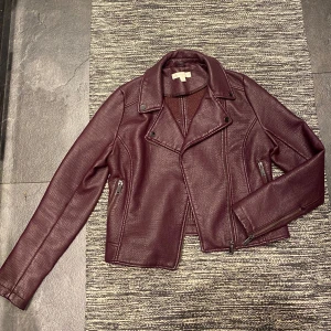 Vinröd skinnjacka från New York & Company - Snygg vinröd skinnjacka från New York & Company. Jackan har dragkedjor vid fickor och ärmslut, bred krage och markerade sömmar. Perfekt för dig som vill sticka ut med en mörk, stilren jacka i skinnimitation!