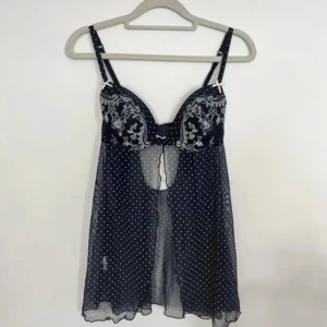 Mörkblå prickig blus med brodyr - Supersöt mörkblå blus i mesh med vita prickar och broderade detaljer på kuporna. Den har tunna axelband, liten rosett framtill och en öppen detalj under bysten. Luftigt och lätt material som ger en romantisk vibe.