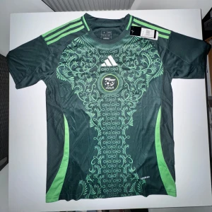  Adidas X Algeriet fotbollströja med mönster - Snygg mörkgrön fotbollströja från Adidas med ljusgröna detaljer och unikt blommigt mönster framtill. STORLEK M! Klassiska Adidas-ränder på axlarna och rund halsringning. Perfekt för dig som vill sticka ut på planen.