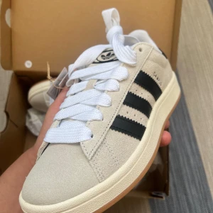 Adidas Campus i beige mocka - Helt nya snygga Adidas Campus sneakers i beige mocka med klassiska svarta ränder på sidorna och vit sula. Skorna har breda vita skosnören och svart Adidas-logga på plösen. Perfekta för dig som gillar retrostil och streetwear.
