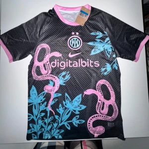 Inter Milan x Nike svart fotbollströja  - Unik Inter Milan fotbollströja från Nike i svart med rosa och blå detaljer. Tröjan har korta ärmar, rosa ormmotiv och blå blommor på framsidan, samt klubbmärke och Nike-logga. Tillverkad i lätt, andningsbart material perfekt för match eller träning.