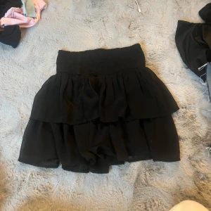 Svart volangkjol med resårmidja - Supersöt svart kortkjol med dubbla volanger och bred smockresår i midjan. Kjolen är luftig och flowy, perfekt för att skapa en chill och trendig look. Passar lika bra till en croppad topp som till en hoodie.