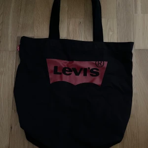 Svart shoppingväska från Levi's - Stor svart shoppingväska från Levi's med klassisk röd logga framtill. Väskan har två breda handtag och är gjord i slitstark bomull, perfekt för att bära med sig allt man behöver. Enkel och stilren design som funkar till det mesta. Använd men i mycket bra skick 