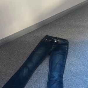 True Religion bootcut jeans mörkblå - Mörkblå jeans från True Religion, modell Becca Mid Rise Bootcut. Snygga kontrastsömmar, bakfickor med lock och knappar. Bootcut passform och midwaist. Lite slitningar längst ner men det är inget som syns.