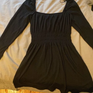 Svart långärmad kortklänning - Supersnygg svart kortklänning med långa ärmar och smockad midja som ger en riktigt fin passform. Fyrkantig urringning och flowy kjol gör den både trendig och bekväm. Perfekt för dig som gillar stilrena och enkla plagg med en twist.