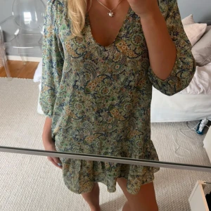 Blommig kortklänning med volang - Supersöt kortklänning med blommigt mönster i grönt, blått och beige. Klänningen har v-ringning, trekvartsärmar och volang nertill. Lätt och flowy modell som är perfekt för varma dagar.