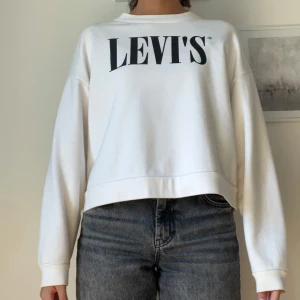 Vit sweatshirt från Levi's, croppad - Vit sweatshirt från Levi's med svart logga framtill och baktill. Modellen är croppad med avslappnad passform och långa ärmar. Tillverkad i mjuk bomull, perfekt för en chill och stilren look.