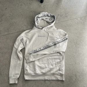 Vit hoodie från GANT med logga - Snygg vit hoodie från GANT med broderad logga på bröstet och GANT-tryck längs ärmarna. Tröjan har huva med snörning och känguruficka framtill. Perfekt för en chill och stilren look.