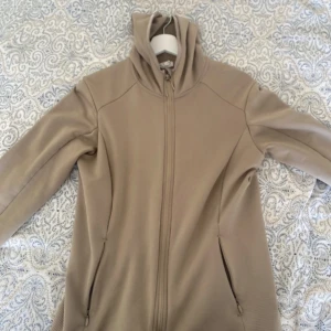 Beige sportig jacka från H&M Sport - Beige sportjacka från H&M Sport i storlek M. Jackan har luva, dragkedja framtill och två fickor med dragkedja. Insidan är fodrad med mjuk fleece för extra komfort. Perfekt för dig som gillar en clean och enkel stil.