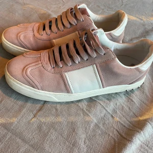 Valentino Garavani sneakers i rosa mocka - Snygga sneakers från Valentino Garavani i ljusrosa mocka och vitt skinn. Skorna har breda snören, vita detaljer och coola nitar runt hälen. Yttersulan har ett unikt camouflagemönster. Perfekta för dig som vill sticka ut med stilrena detaljer. Hör av dig vid frågor 