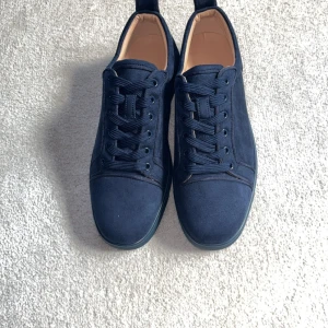 Mörkblå Christian Louboutin sneakers - Snygga mörkblå sneakers från Christian Louboutin i mocka med ikoniska röda sulor. Skorna har rund tå, snörning och diskret branding på sulan. Perfekta för dig som vill sticka ut med exklusiv stil och lyxiga detaljer.