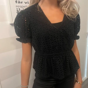 Svart blus med puffärm och broderi - Supersöt svart blus med v-ringning, puffiga korta ärmar och volang nertill. Blusen har broderade hål och markerad midja som ger en snygg siluett. Perfekt att styla med jeans eller kjol för en trendig look.