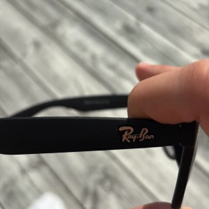 Svarta Ray-Ban Wayfarer solglasögon - Klassiska svarta Ray-Ban Wayfarer solglasögon med mattsvart båge och mörka glas. Snygg och tidlös design som passar till allt. Modellnummer GR-037, storlek 60-16-139. Ray-Ban-logga på skalmen och linsen för extra premiumkänsla. 1|1 modell