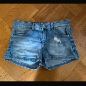 Blå jeansshorts med slitningar - Snygga blå jeansshorts med slitna detaljer framtill och klassisk femficksmodell. Shortsen har bälteshällor, knapp och dragkedja i midjan samt en avslappnad passform. Perfekta för varma dagar när du vill ha en chill och trendig look.