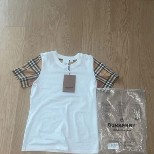Vit Burberry t-shirt med rutiga ärmar - Snygg vit t-shirt från Burberry med klassiska rutiga ärmar i beige, svart, rött och vitt. T-shirten har rund halsringning och är tillverkad i mjuk bomull. Perfekt för dig som vill ha en stilren look med en touch av lyx.