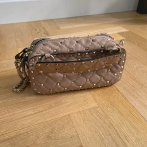 Beige quiltad axelväska Valentino - Säljer en beige quiltad axelväska från Valentino Garavani med guldiga nitar och kedjerem. Väskan är i skinn och har dragkedja upptill samt ett snyggt rött innerfoder. Perfekt för dig som gillar lyxiga detaljer och stilren design.Såå fin! Har med äkthetsverifikationen signerad från Vinted! Säljer då jag ska köpa en annan🥂🫶🏼 Finaste väskan och självklart äkta🥰🥰