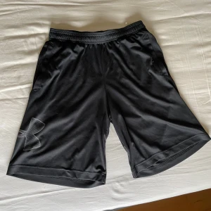 Svarta träningsshorts från Under Armour - Svarta träningsshorts från Under Armour med elastisk midja och två sidofickor. Stort logotryck på vänster ben. Tillverkade i ett lätt och svalt polyestermaterial som passar perfekt för sport och träning.