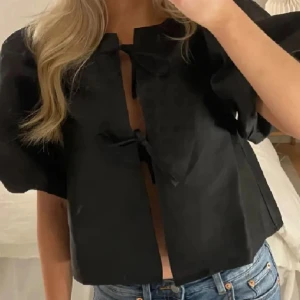 Svart blus med puffärm och knyt - Säljer en svart blus med korta puffärmar och två knyt framtill. Blusen är croppad och har en luftig passform som ger en cool och trendig vibe. Perfekt att styla med jeans eller kjol för en snygg look.