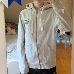 Ljusgrå hoodie från Dobber - Säljer en ljusgrå hoodie från Dobber med dragkedja och huva. Tröjan har vita snören, två fickor framtill och broderad logga på bröstet. Perfekt för chill dagar och enkel att matcha med det mesta.
