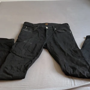 Svarta jeans från Lee - Säljer ett par svarta jeans från Lee med klassisk femficksdesign och raka ben. Jeansen har Lee-logga på bakfickan och svart patch i midjan. Snygg och enkel modell som passar till det mesta. Materialet är jeans och färgen är helsvart. Hör gärna av er vid minsta lilla fråga 