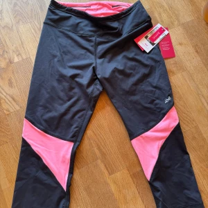 Svarta och rosa tights från Röhnisch - Snygga svarta, korta tights från Röhnisch med rosa detaljer i modellen Shape Aine Capri. De har hög midja och croppad längd, perfekt för träning. Materialet är stretchigt syntet som ger en tight passform och bra stöd. Superskön känsla och cool design! Ordinarie pris 599kr