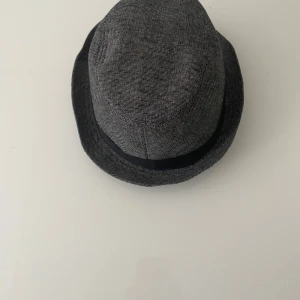Grå rutig fedora hatt M - Snygg grå fedora hatt i rutigt mönster med svart band runt kullen. Klassisk form med smal brätte och stilren look, perfekt för dig som vill sticka ut med en cool accessoar. Tillverkad i ett mjukt tyg som känns bekvämt på huvudet.