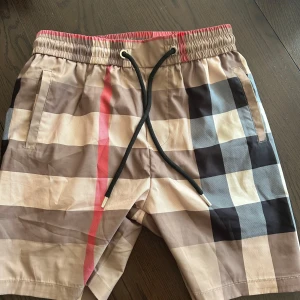 Rutig Burberry shorts med snörning - Snygga shorts från Burberry med klassiskt rutigt mönster i beige, svart, grått och rött. De har elastisk midja med svart snörning och fickor på sidorna samt bak. Perfekta för sommaren och ger en clean, lyxig vibe.