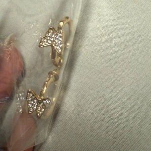 Guldiga örhängen med strassfjärilar - Snygga guldiga örhängen i creolmodell med oregelbunden, bambuliknande form. Varje örhänge har en liten fjäril framtill som är täckt av glittrande strass-stenar. Perfekta för dig som vill ha något extra och trendigt i öronen.