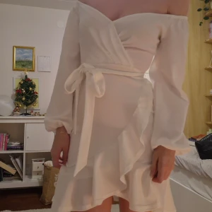 Vit omlottklänning - Elegant vit omlottklänning med offshoulder-design och volangdetaljer. Klänningen har långa ärmar och ett knytband i midjan för en smickrande passform. Aldrig använd, passar s också. 