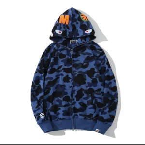 Blå camo hoodie från Bape - Säljer en blå camo hoodie från Bape med dragkedja och huva. Hoodien har svarta och mörkblå camouflage-mönster, broderade detaljer och ögon på huvan. Perfekt för dig som vill sticka ut med streetwear-stil.