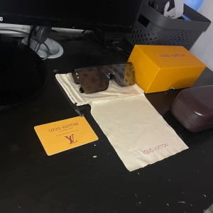 Louis Vuitton monogram solglasögon - Snygga solglasögon från Louis Vuitton med rektangulära, stora svarta bågar och bruna linser med LV-monogram över hela glaset. Glasögonen har en exklusiv vibe och levereras med originalfodral, box och certifikat. Perfekt accessoar för dig som vill sticka ut.