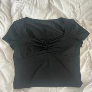 Svart croppad topp med rynk framtill - Snygg svart croppad topp med korta ärmar och rynkdetalj vid bysten. Toppen har en fyrkantig ringning och är figurnära, perfekt att styla med höga jeans eller kjol. Mjuk och stretchig känsla.