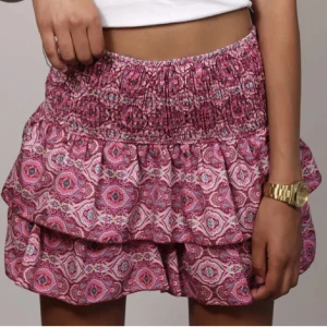 Rosa mönstrad volangkjol - Meet me there kjol 💕Supersöt kort kjol i rosa med volanger och elastisk midja. Kjolen har ett detaljerat mönster i vitt och lila, perfekt för en färgglad look. Lätt och luftig känsla, passar perfekt till varma dagar.