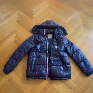 Mörkblå Moncler dunjacka med huva - Snygg mörkblå dunjacka från Moncler med glansig finish och avtagbar huva. Jackan har dragkedja med röd, vit och blå detalj, flera fickor och klassisk Moncler-logga på bröstet. Perfekt för kalla dagar när du vill hålla stilen.