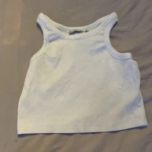 Vit ribbad linnetopp - Säljer en enkel och fräsch vit ribbad linnetopp med breda axelband. Toppen är croppad och har en klassisk, stilren look som passar perfekt till jeans eller kjol. Materialet är mjukt och stretchigt för en bekväm passform.