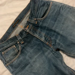 Blå jeans från Nudie Jeans Co - Snygga blå jeans från Nudie Jeans Co med klassisk femficksdesign och orangea kontrastsömmar. Jeansen har raka ben och coola orangea render på  bakfickor med vågigt mönster. Tillverkade i slitstarkt bomullsjeansmaterial och passar perfekt till en Grinch Steel