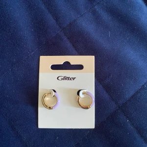 Lila och guldiga hoops från Glitter - Säljer ett par snygga små hoops från Glitter i guld med lila detalj. Perfekta för att ge din look en färgklick! Runda örhängen med stilren design som passar till många outfits.