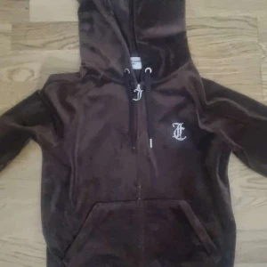Brun velour hoodie Juicy Couture S - Mörkbrun velour hoodie från Juicy Couture med dragkedja, huva och känguruficka. Stora strasstenar bildar 'Juicy' på ryggen och ett JC-märke framtill. Perfekt för dig som gillar 2000-talsvibbar och comfy stil.