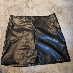 Svart kort kjol i skinnimitation - Snygg svart minikjol i skinnimitation med stilren design och diskret söm framtill. Kjolen har en rak passform och dragkedja i sidan. Perfekt för dig som vill ha en edgy vibe till outfiten.