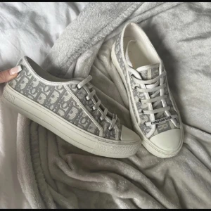 Grå Dior sneakers med monogram - Snygga sneakers från Dior i grått och vitt med ikoniskt monogrammönster. Skorna har snörning, vit platt sula med svart stjärndetalj undertill och rund tå. Ovandelen är i textil med tydlig Dior-logga framtill och på sidan. Perfekta för dig som gillar lyxig streetstyle.